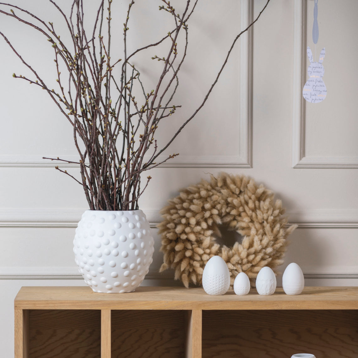 Decorative table with four white porcelain embossed eggs, textured white vase, and branches on a wooden surface.