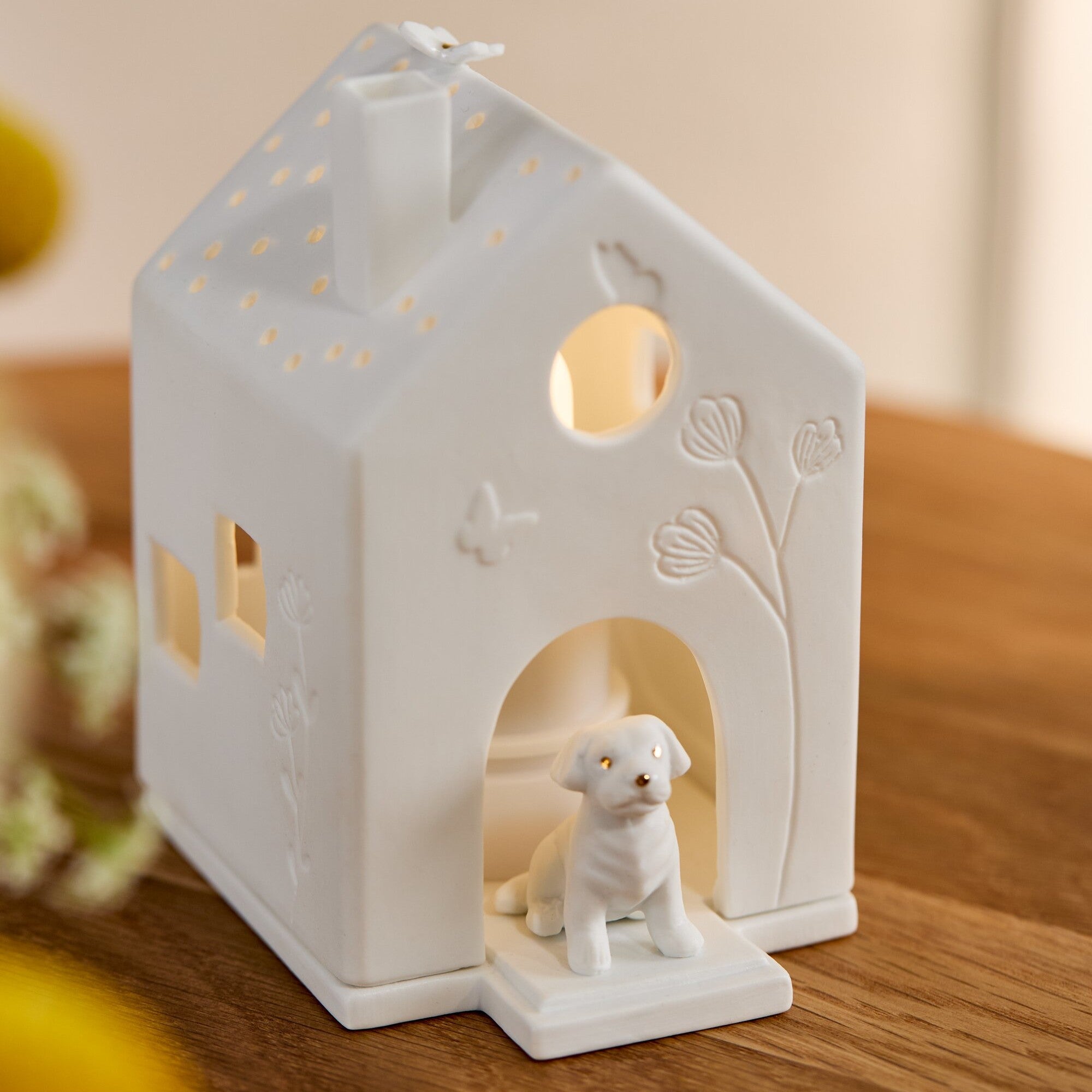 A white porcelain dog-shaped house candle holder with a small white dog figurine sitting in the doorway, placed on a wooden surface.