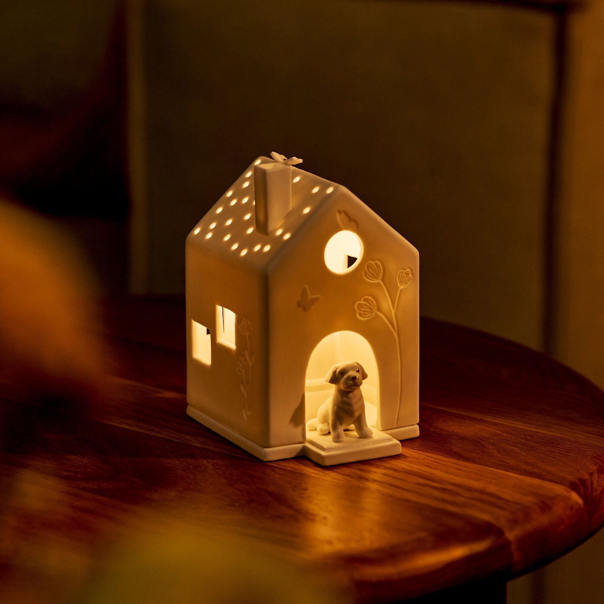 A warm white porcelain house-shaped night light with a small dog figure sitting in the doorway, placed on a wooden table in a dimly lit room.