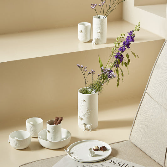 White porcelain stores vases, bowls and plates with purple flowers on a beige surface.