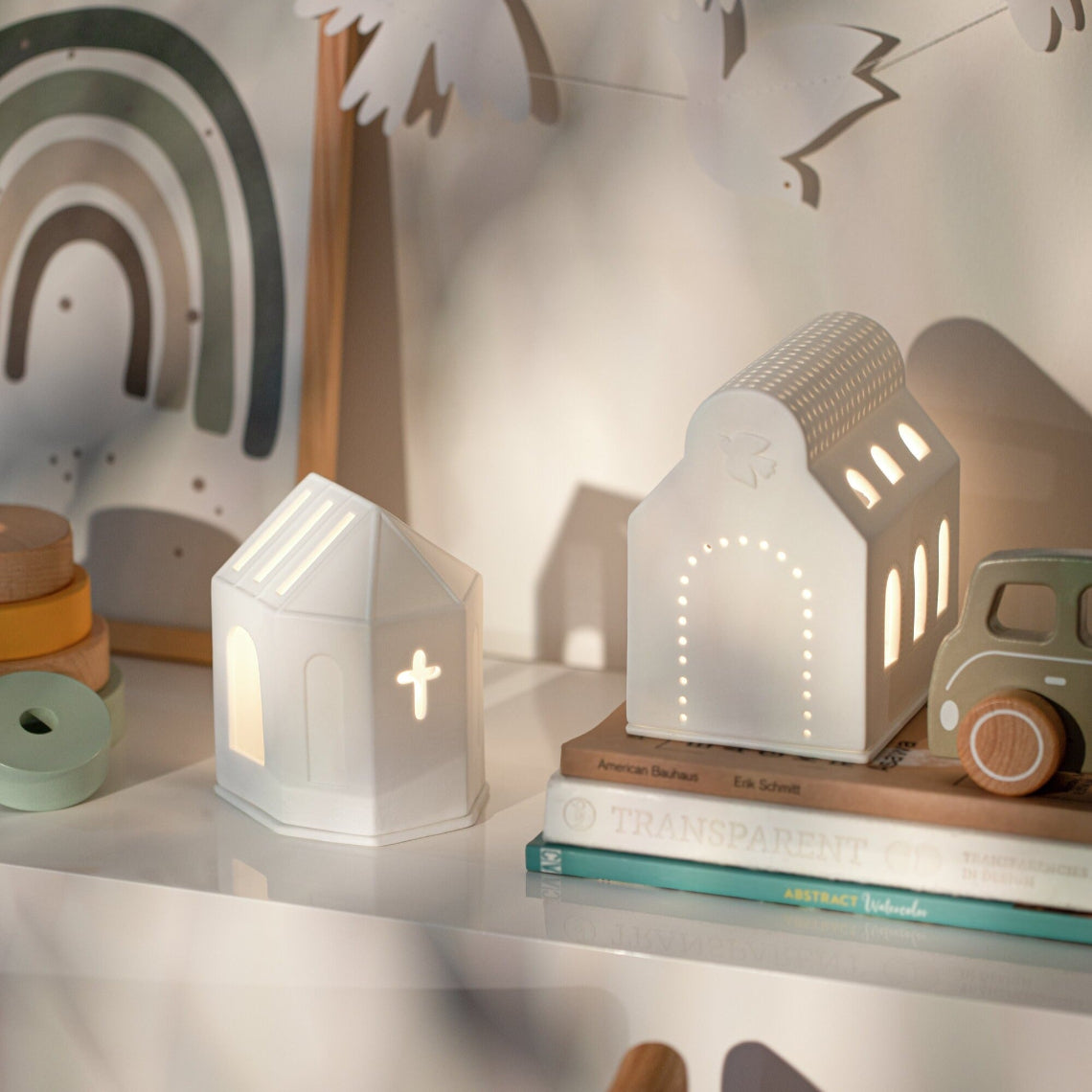 House-shaped tealight holders on a shelf with books and toys in a cozy room setting.