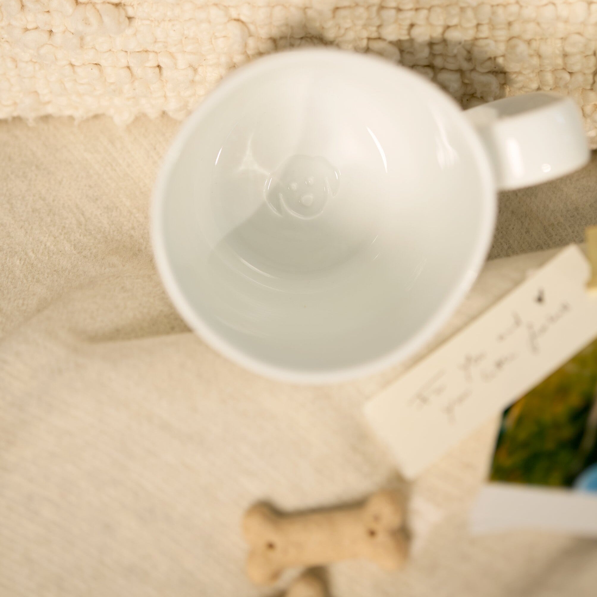 A white porcelain mug shown from a top-down angle on a textured cream fabric surface, with a small bone-shaped prop and a blurred tag nearby.