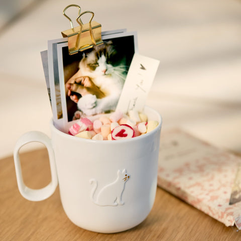 A white porcelain mug with a cat silhouette on the front, filled with pastel candy and holding printed cards and photos with a gold binder clip, set on a wooden surface.