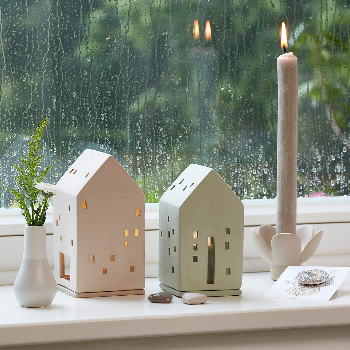 Two house-shaped candle holders on a windowsill with a candle and plant in the background.