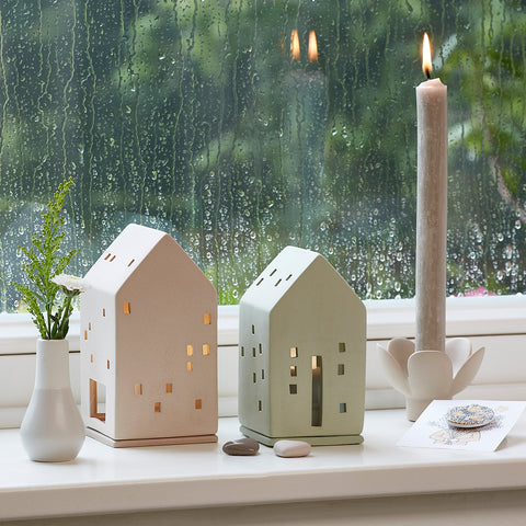 Two house-shaped candle holders on a windowsill with a candle and plant in the background.