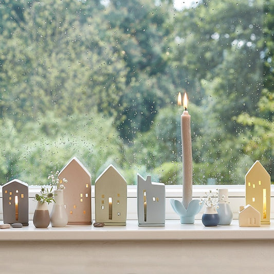 Decorative stoneware houses on a windowsill with a blurred green outdoor background, vases and candles.